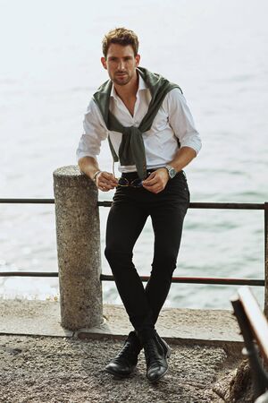 Stylish man wearing white shirt with tied sweater on shoulders over lake backgroundの写真素材