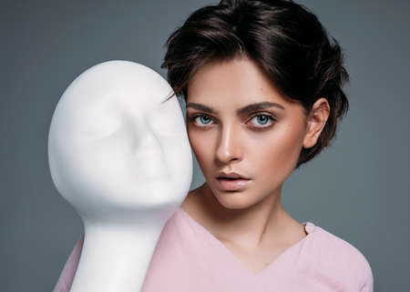 Portrait of young brunette woman with white mannequin face isolated on gray backgroundの写真素材