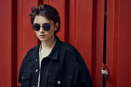 Portrait of a young woman in sunglasses wear black denim jacket over red backgroundの写真素材