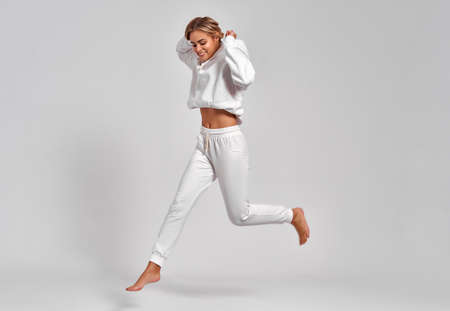 Beautiful blonde woman jumping in the air in white tracksuit isolated on gray backgroundの写真素材