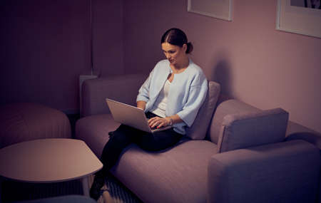 Late At Night Beautiful Woman Sitting on Her Sofa in the Living Room with a Laptop on Her Kneesの写真素材