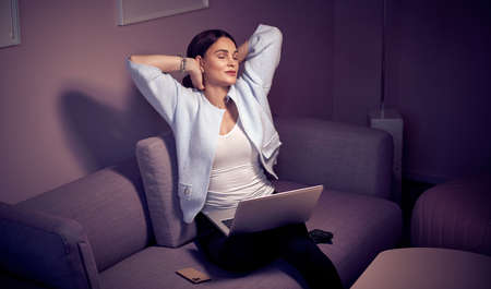 Late At Night Beautiful Woman Sitting on Her Sofa in the Living Room with a Laptop on Her Kneesの写真素材
