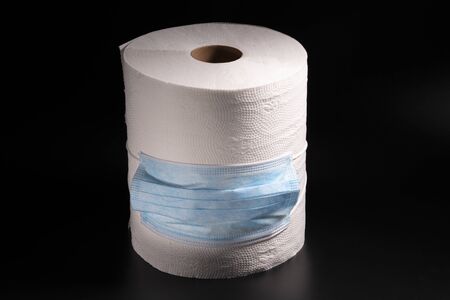 A large roll of toilet paper or paper towels with a medical mask on a black background, close-upの写真素材
