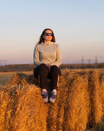 A girl with long hair sits on a haystack at sunset wearing glasses.