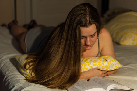 A girl lies on a bed and reads a book, dimmed light