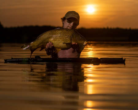 A man in the water holds a large carp. Beautiful sunset on the water.の写真素材