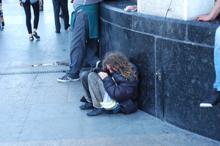 The slovenly homeless man sits on the ground near the wall, waiting for alms.のeditorial素材