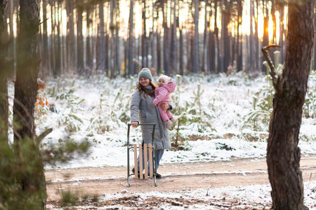 Mother walking in snowy forest with daughter in pink snowsuit and sledの写真素材