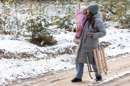 Mother walking in snowy forest with daughter in pink snowsuit and sledの写真素材