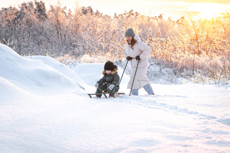A young mother takes her son sledding in a snowy area against the backdrop of a sunset - a concept...