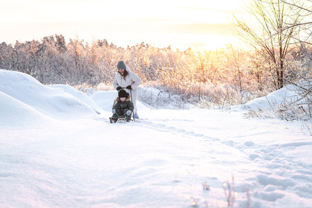 A young mother takes her son sledding in a snowy area against the backdrop of a sunset - a concept...