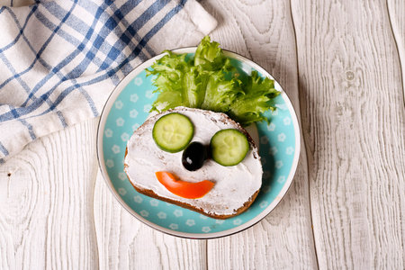Kids meal, breakfast or lunch for children. Colorful funny food face with healthy vegetables, cheese spread and green salad on a plate. Concept of healthy food, healthy breakfast. Top view, closeup, horizontalの写真素材