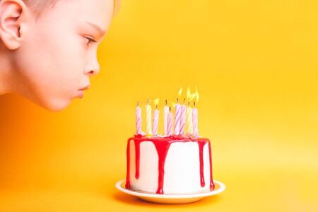 Boy blows out candles on a white cake with red icing on an orange background. Birthday concept.の写真素材