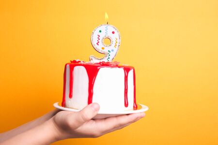 White cream cake with red icing and a candle in children's hands on an orange background. Birthday concept.の写真素材