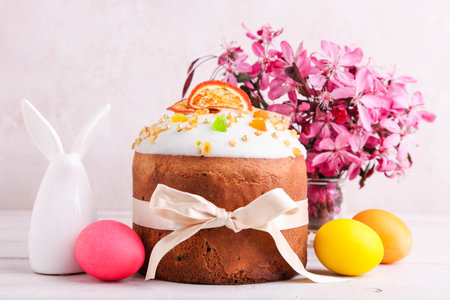 Easter cake with colored eggs and bouquet of pink flowers on a white wooden background.
