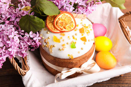 Easter cake with colored eggs and branch of lilac in wicker basket on brown wooden background.の写真素材