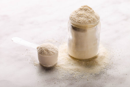 White protein powder in jar with measuring spoon on a marble background.