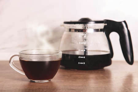 Hot coffee in glass cup and jug of coffee maker on wooden kitchen countertop.の写真素材