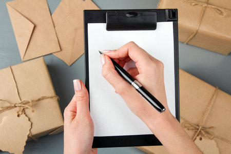 woman's hand writes on paper. Parcels in craft paper and envelopes on gray wooden background....