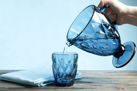 Woman's hand pouring water from jug into glass on blue background.