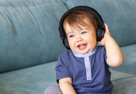 Adorable little baby boy listening to music in headphones on his head sitting on a blue sofa at home and smiling with his first eight teeth, musical education concept,の写真素材