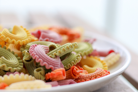 Colorful green, yellow, white, orange and pink italian crown pasta on white plate staying on wooden table, close-up. Multicolor pasta texture.  Front focus, copy spaceの写真素材