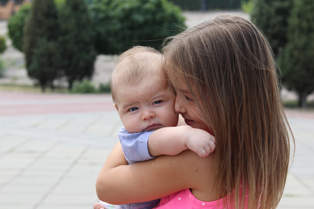 Young girl holding her newborn brother in her arms and looking at him with tenderness, outdoorsの写真素材