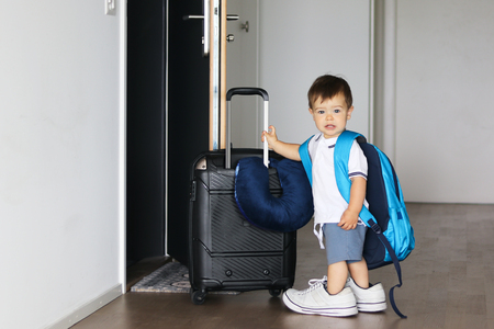 Cute little baby boy in fathers shoes and with big backpack holding suitcase staying near open door ready to travel leaving home. Traveling with child conceptの写真素材