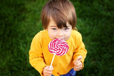 View from above at little boy eating big red lollipop outdoors on green grass looking up to camera. Sweet tooth. Carefree childhood.の写真素材