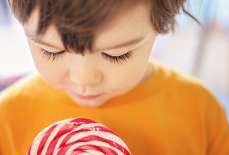 Close up face of little boy with long eyelashes looking at striped lollipop. Sweet childhood. Selective focus on eyelashes. Foretasteの写真素材