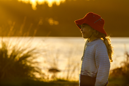 close up of a four year old child wearing a bright red hat  standing on the shore of a lake at sunsetの写真素材