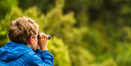close up of a young boy in a blue coat looking through binoculars at birds with a blurred green backgroundの写真素材