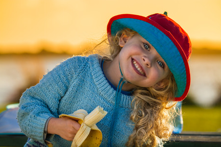 close up of a smiling child wearing a hat and holding a half eaten banana at sunsetの写真素材
