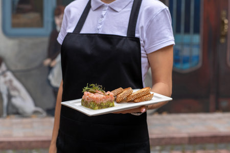 a girl in a black apron and a white bozka is holding in front of her a salmon avocado and rucola...
