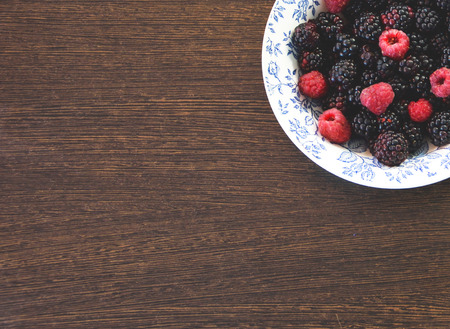 Fresh sweet blackberries and raspberries in a white bowl with a blue pattern against the dark wooden surface of the table. A place for a label.の写真素材