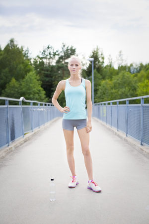 Beautiful and fit young blonde woman taking a break after jogging. Standing with her hand on her hip on a bridge.の写真素材