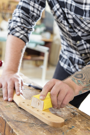 Craftsman using sanding paper on a guitar neck in a workshop for wood. Hard working man with tattoo working with musical instruments.の写真素材