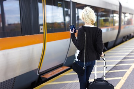 Young woman with mobile phone at outdoor train terminal with a wheeled suitcase. Traveling with public transport.