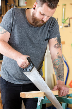 Hardworking carpenter sawing with a hand saw in work shop for woodwork. Man with tattoo and beard.の写真素材