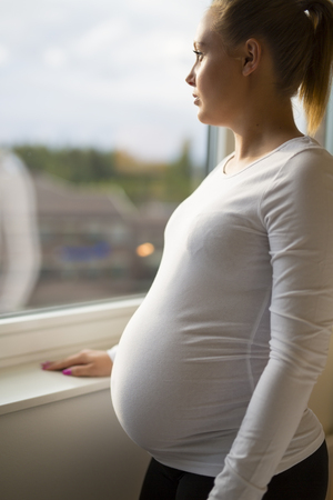 Young thoughtful pregnant woman looks out the window home. Holding a hand on the tummy. Mental health and pregnancy.の写真素材