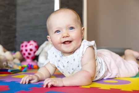 Happy baby girl with blue eyes playing on the floor with toys on a playmat. A cute young child plays and smiles.の写真素材
