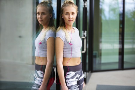 Portrait of a beautiful blonde woman in sportswear who takes a break after workout. Stands in urban city environment in tights and crop top. Reflection in window.の写真素材