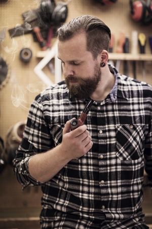 Environmental portrait of a young adult craftsman sitting and smoking a pipe tobacco in a workshop for wood work and construction.の写真素材