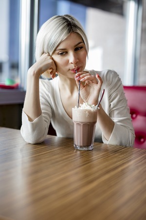 Beautiful young woman drinking hot chocolate with cream at table in cafeの写真素材
