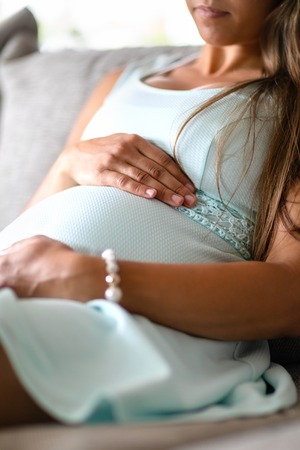 Close-up of pregnant woman sitting in sofa with her hands at bellyの写真素材