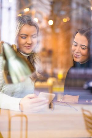 Two Female Friends Looking At Smartphone In Cafe seen through windowの写真素材