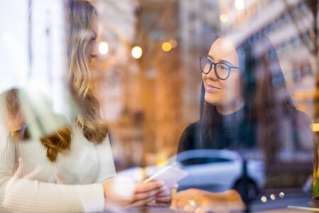 Two Young Women Talking At Cafe in City Seen Through Windowの写真素材