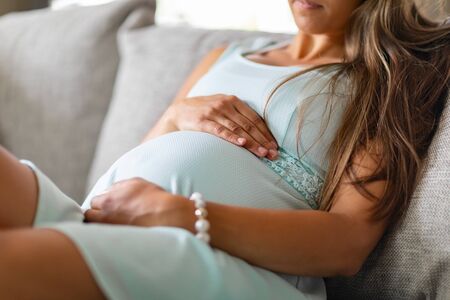 Mid-section of pregnant woman sitting in sofa with her hands at bellyの写真素材