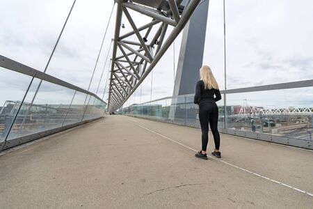Rear view of blonde fitness woman runner standing on bridge in modern looking cityの写真素材
