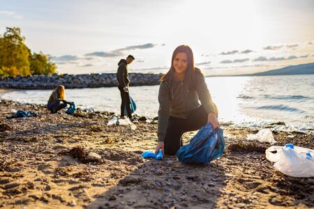 Smiling young woman cleaning beach with volunteers during sunsetの写真素材